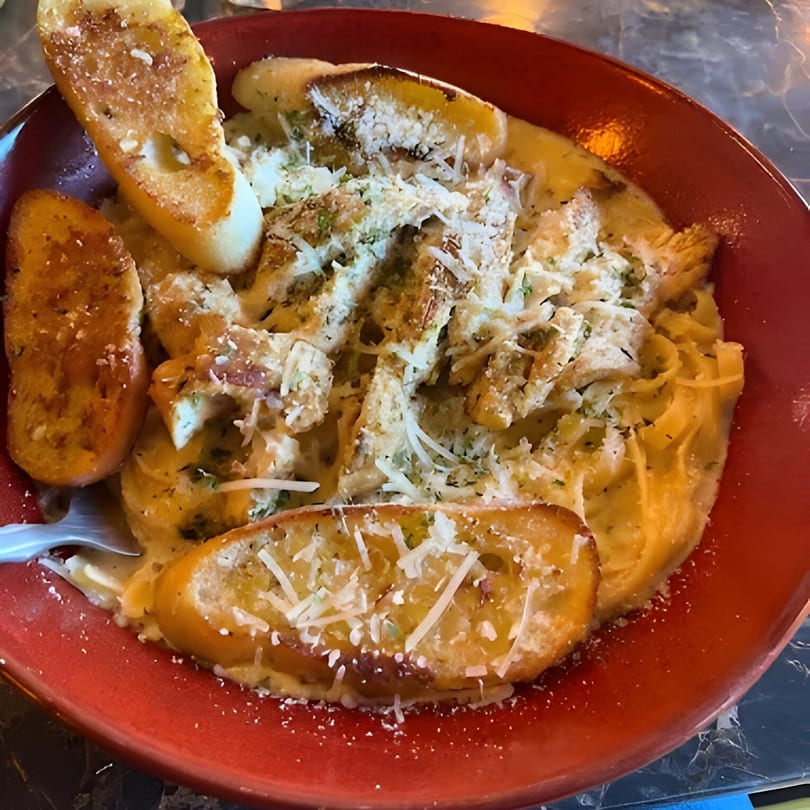 Creamy pasta with chicken and garlic bread, topped with parmesan cheese and herbs in a red bowl.