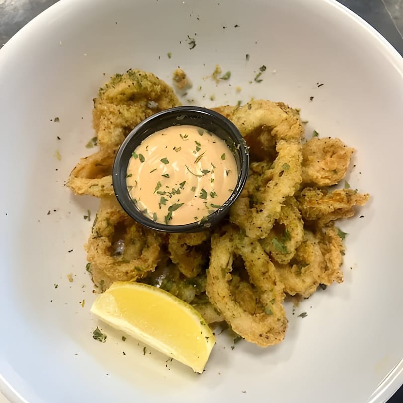 Crispy fried calamari rings with a side of creamy sauce and a lemon wedge in a white bowl.